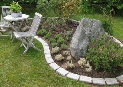 En trädgård med en stensättning runt en rabatt och ett bord och två stolar